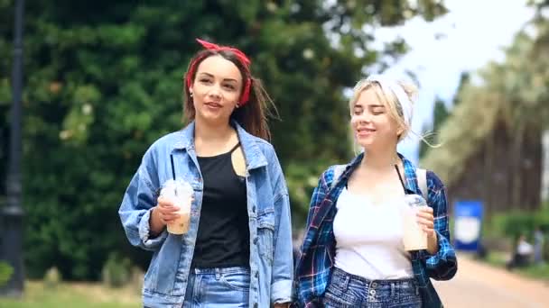 Две девушки прогуливаются по городской улице и пьют молочный коктейль из одноразовой чашки — стоковое видео