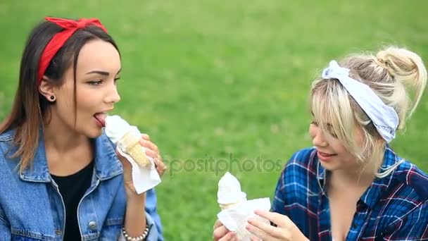 Zwei gut gelaunte Freundinnen sitzen auf dem Rasen im Stadtpark und essen Eis — Stockvideo