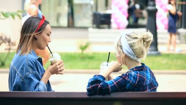 Dos amigos se sientan en un banco en el parque de la ciudad y beben un batido de una taza desechable — Vídeos de Stock