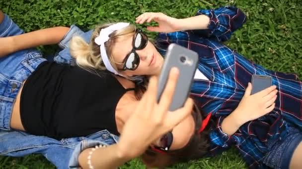 Two cheerful cute girlfriends do selfie on the phone sitting on the green glade of the city park — Stock Video