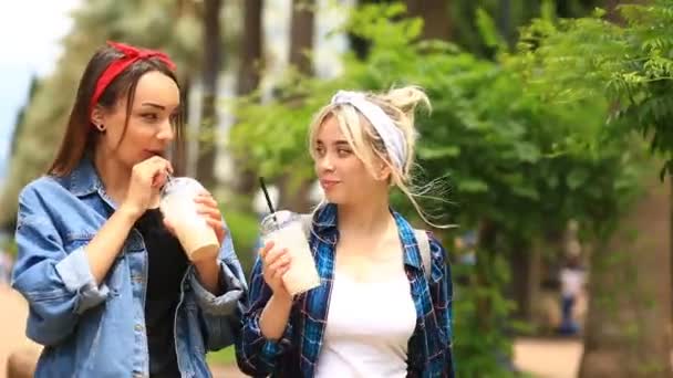 Zwei Freundinnen schlendern die Straße entlang und trinken einen Milchshake aus einem Einwegbecher — Stockvideo