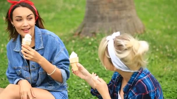 Dos novias alegres sentadas en el césped en el parque de la ciudad y comiendo helado — Vídeos de Stock