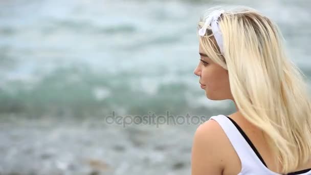 Молодая женщина сидит на пляже, мило улыбается и смотрит в камеру на фоне морских волн — стоковое видео