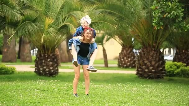 背景にはヤシの木と緑の芝生の公園で 2 つの面白い女の子のガール フレンドはしゃぐ。1 つは別の背面にジャンプします。 — ストック動画