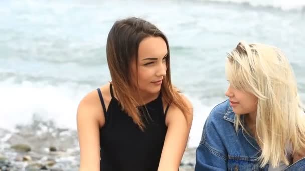 Dos hermosas amigas sentadas en la playa contra las olas del mar y mirando a la cámara — Vídeos de Stock