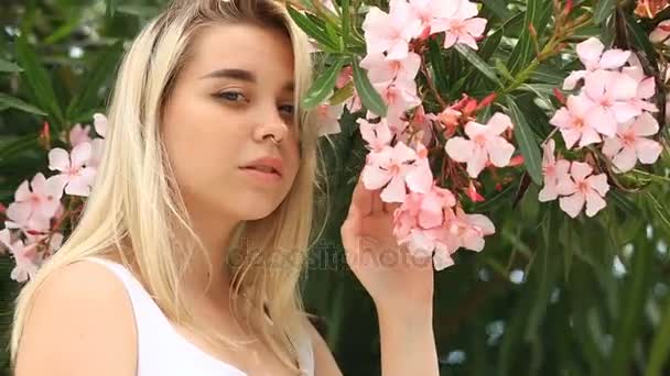 Mooie jonge blonde vrouw glimlacht lief en kijkend naar de camera op een achtergrond van roze bloeiende boom bloemen — Stockvideo