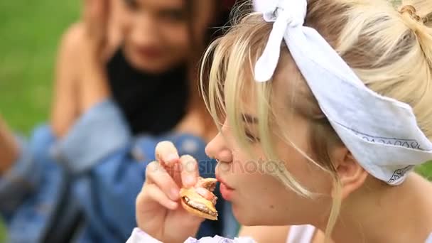 Dos amigas comen hamburguesas, papas fritas en un césped verde en el jardín de la ciudad. comida rápida — Vídeos de Stock