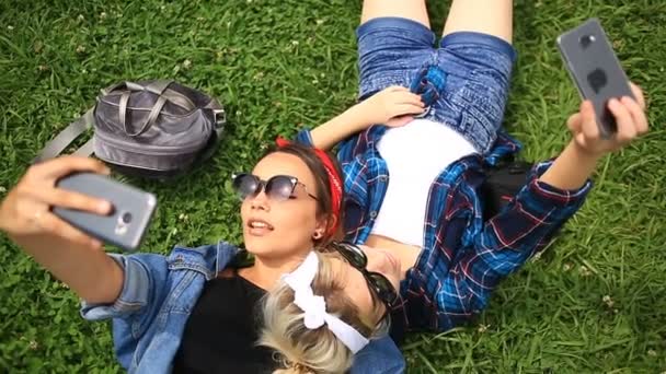 Two cheerful cute girlfriends do selfie on the phone sitting on the green glade of the city park — Stock Video