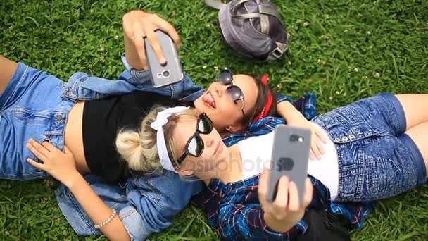 Two cheerful cute girlfriends do selfie on the phone sitting on the green glade of the city park — Stock Video