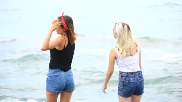 Duas namoradas fofas alegres estão se divertindo na praia de pé no mar, jogar pedras na água — Vídeo de Stock