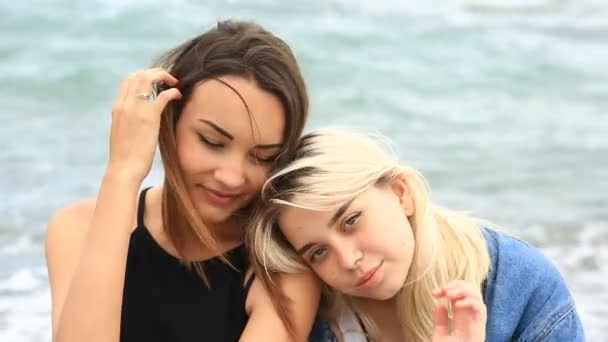 Dos hermosas amigas sentadas en la playa contra las olas del mar y mirando a la cámara — Vídeos de Stock