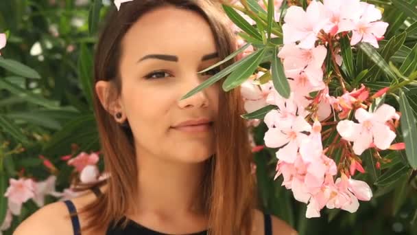 Mooie jonge brunette glimlachend schattig en kijkend naar de camera op een achtergrond van roze bloeiende boom bloemen — Stockvideo