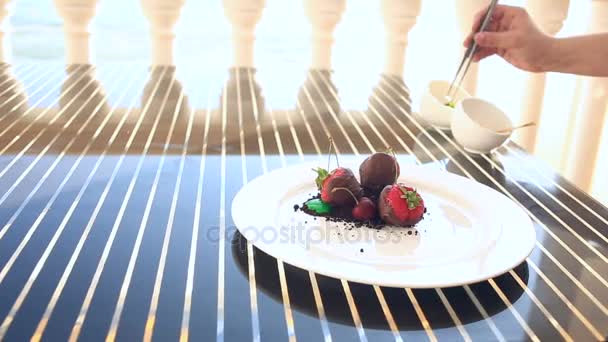 Dit is een gastronomische schotel. Close-up. Dessert, aardbei in de chocolade. Met een glas witte wijn. Seaside restaurant met uitzicht op zee. Vrouwelijke hand neemt bessen met een vork — Stockvideo
