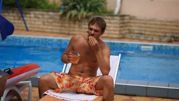 П'яний чоловік у шортах п'є пиво біля басейну, курить цигарку — стокове відео