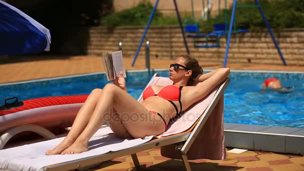 Молодая, элегантная женщина читает книгу на шезлонге у бассейна — стоковое видео