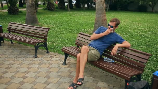 Человек сидит на скамейке в городском парке и измучен жарой. . — стоковое видео