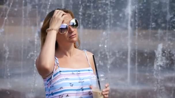 Une femme blonde aux lunettes de soleil se tient à une fontaine par temps chaud avec un cocktail laiteux. au ralenti — Video