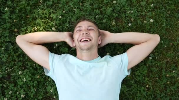 Joven chico guapo yace en la hierba en el parque de verano y se ríe mirando la cámara. Movimiento lento — Vídeos de Stock