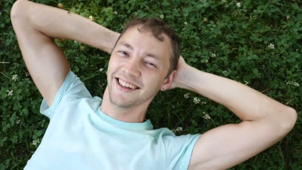 젊은 잘생긴 남자 여름 공원에서 잔디에 속이 고 카메라를 보고 웃는 다. 슬로우 모션 — 비디오