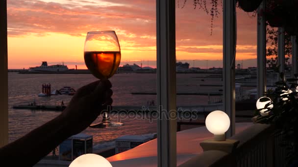 Un hombre sostiene un vaso de alcohol contra la puesta de sol sobre el mar. Primer plano. Movimiento lento — Vídeo de stock