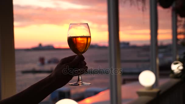 Um homem está segurando um copo de álcool contra o pôr do sol sobre o mar. close-up. Movimento lento — Vídeo de Stock