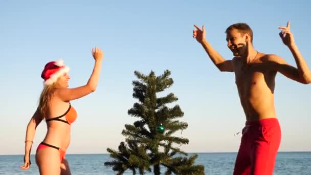 Par kille och tjej i santa hatt hälsade det nya året och jul på stranden. Att dekorera en julgran på en tropisk strand. Slow motion — Stockvideo