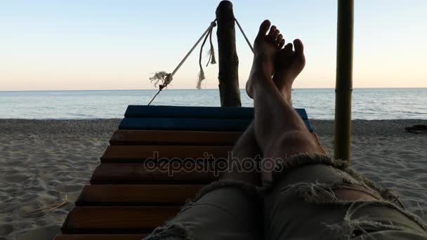 Lábát lóbálva egy függőágyban, pihennek a tengerparton naplementekor. lassú mozgás — Stock videók