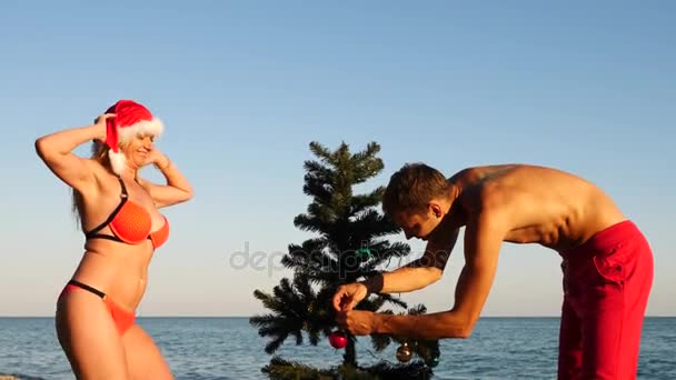 Casal cara e menina em Papai Noel cumprimentou o ano novo e Natal na praia. Decorar uma árvore de Natal em uma praia tropical. Movimento lento — Vídeo de Stock