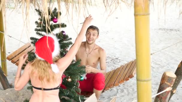 Casal cara e menina em Papai Noel cumprimentou o ano novo e Natal na praia. Decorar uma árvore de Natal em uma praia tropical. Movimento lento — Vídeo de Stock