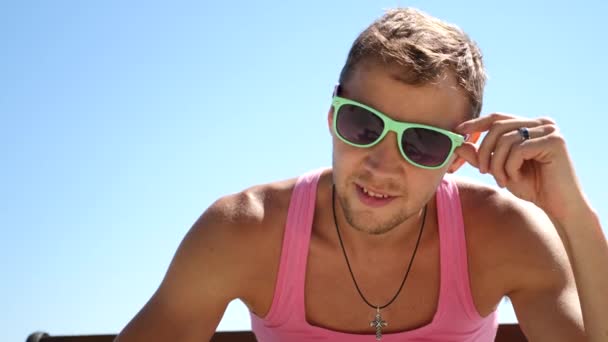 Guapo chico de moda con gafas de sol en una camiseta rosa mira a la cámara y de repente apunta a la cámara — Vídeos de Stock