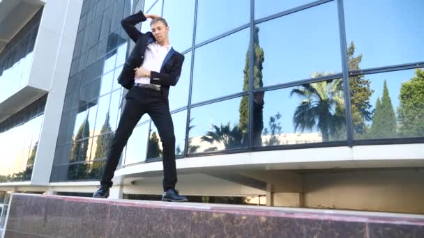 A young happy businessman in a business suit dancing modern ballet and wacking against the backdrop of a business center building. Slow-mo — Stock Video