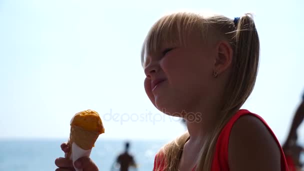 Menina loira comendo sorvete de frutas na praia em um dia ensolarado brilhante. 4K. Movimento lento . — Vídeo de Stock