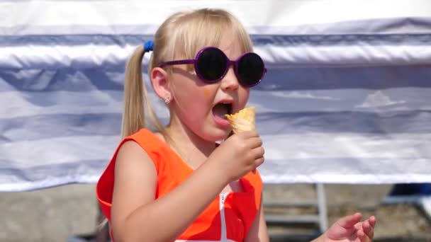 Menina loira comendo sorvete de frutas na praia em um dia ensolarado brilhante. 4K. Movimento lento . — Vídeo de Stock