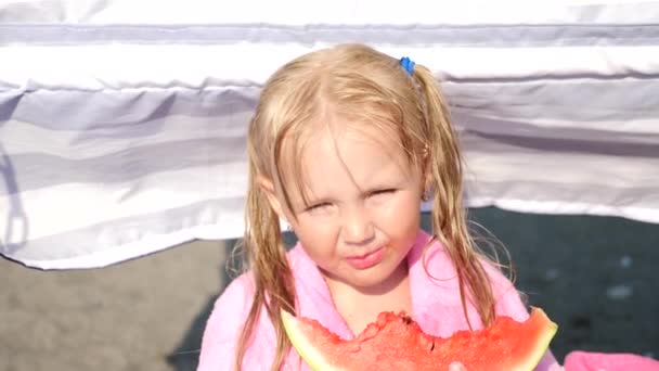 Маленька блондинка в сонцезахисних окулярах їсть фрукти на пляжі. 4K. Повільний рух . — стокове відео