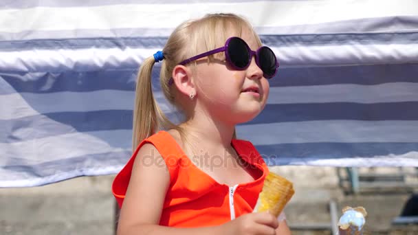 Menina loira comendo sorvete de frutas na praia em um dia ensolarado brilhante. 4K. Movimento lento . — Vídeo de Stock