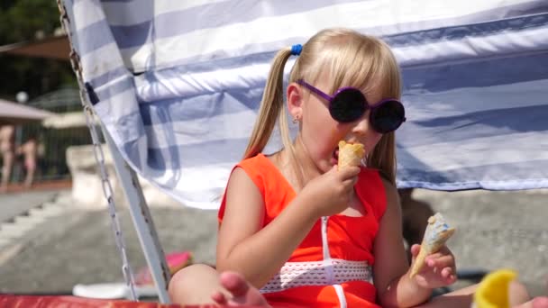 Niña rubia comiendo helado de frutas en la playa en un día soleado brillante. 4K. Movimiento lento . — Vídeos de Stock