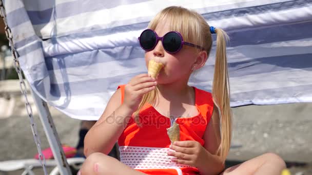 Menina loira comendo sorvete de frutas na praia em um dia ensolarado brilhante. 4K. Movimento lento . — Vídeo de Stock