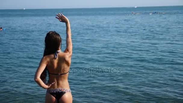 Una chica sexy en un bikini salpica en el mar, de pie con la espalda a la cámara, su figura delgada y las nalgas elásticas son visibles. cámara lenta, 4k . — Vídeo de stock