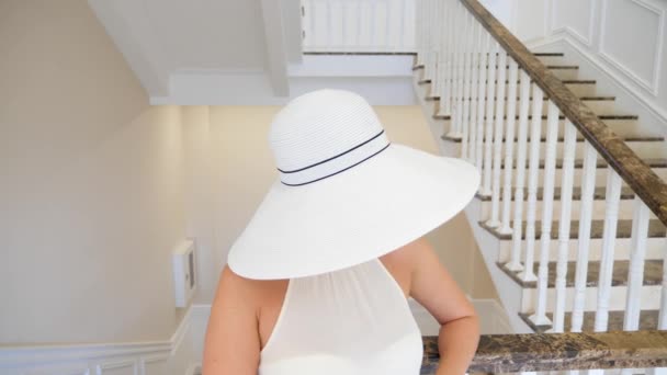 Una mujer con un gran sombrero blanco baja por una gran escalera blanca. 4k. Movimiento lento — Vídeo de stock