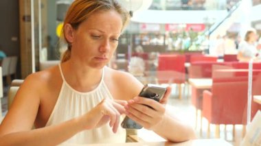 Sarışın bir kadın bir sıcak yaz gününde bir kafede oturan ve üzerinde telefon gülümseyen bir iletide metin yazarak. 4 k. Slow-motion