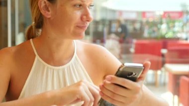 Sarışın bir kadın bir sıcak yaz gününde bir kafede oturan ve üzerinde telefon gülümseyen bir iletide metin yazarak. 4 k. Slow-motion