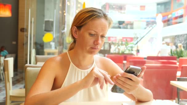 Une femme blonde est assise dans un café par une chaude journée d'été et tape du texte dans un message au téléphone en souriant. 4 k. Mouvement lent — Video