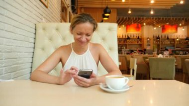 Sarışın bir kadın bir sıcak yaz gününde bir kafede oturan ve üzerinde telefon gülümseyen bir iletide metin yazarak. 4 k. Slow-motion