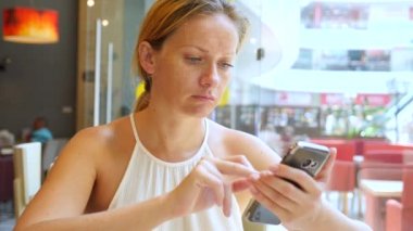 Sarışın bir kadın bir sıcak yaz gününde bir kafede oturan ve üzerinde telefon gülümseyen bir iletide metin yazarak. 4 k. Slow-motion