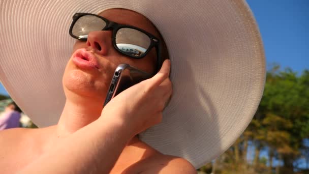 Mujer 4K en sombrero blanco de ala ancha y gafas de sol en la playa usando su teléfono, en cámara lenta . — Vídeo de stock