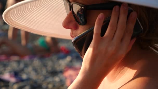 Mujer 4K en sombrero blanco de ala ancha y gafas de sol en la playa usando su teléfono, en cámara lenta . — Vídeos de Stock