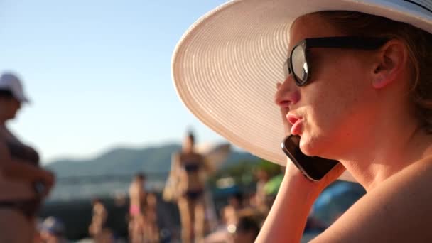 Mujer 4K en sombrero blanco de ala ancha y gafas de sol en la playa usando su teléfono, en cámara lenta . — Vídeo de stock