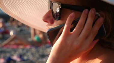 4 k kadında geniş ekledi beyaz şapka ve güneş gözlüğü telefonunu, ağır çekimde kullanarak sahilde.