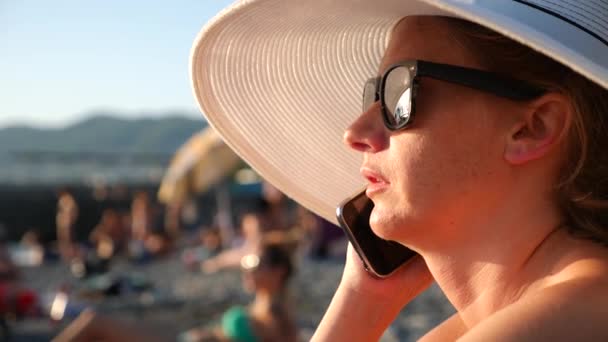 4 k žena v bílém klobouku se širokou krempou a sluneční brýle na pláži pomocí její telefon, zpomalené. — Stock video