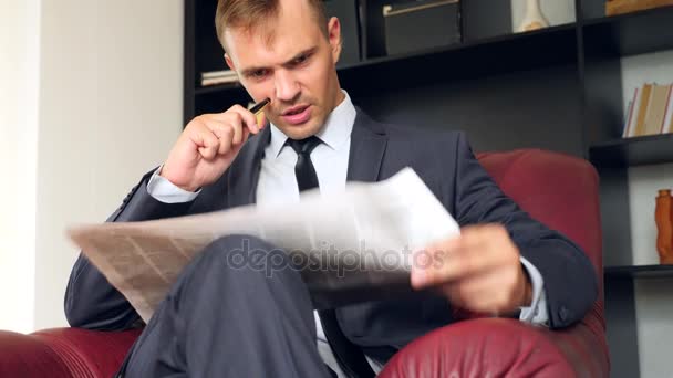 El hombre de negocios está sentado en una silla y leyendo un periódico. Es infeliz y expresivo. 4. Movimiento lento . — Vídeos de Stock